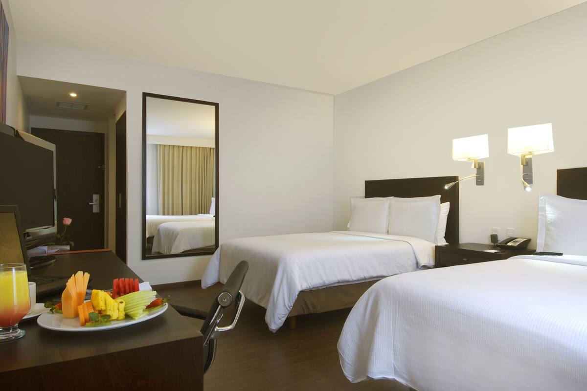 A hotel room with two beds, a desk with a laptop, a plate of fruit, and a mirror.