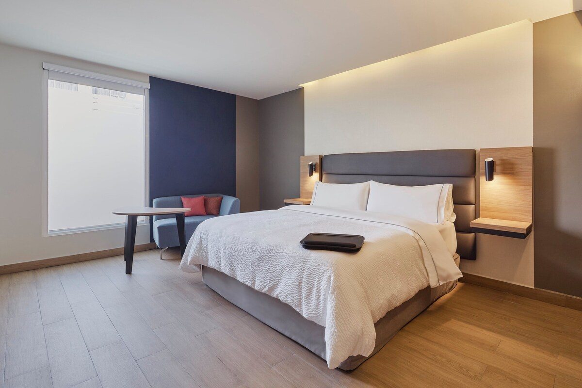 A modern hotel room with a large bed, bedside tables, a small table, and a window.