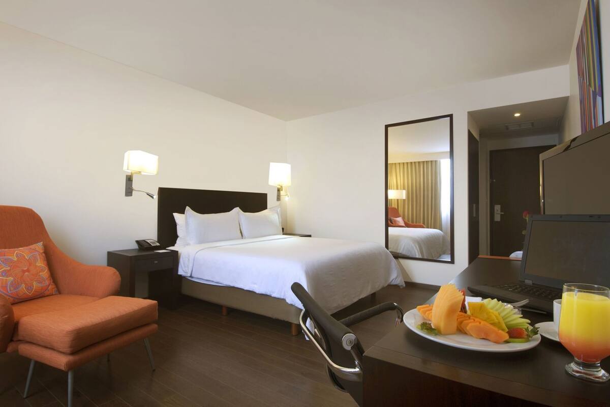 A hotel room with a bed, a desk with a laptop, a chair, a dining table with fruit and juice, and a mirror.