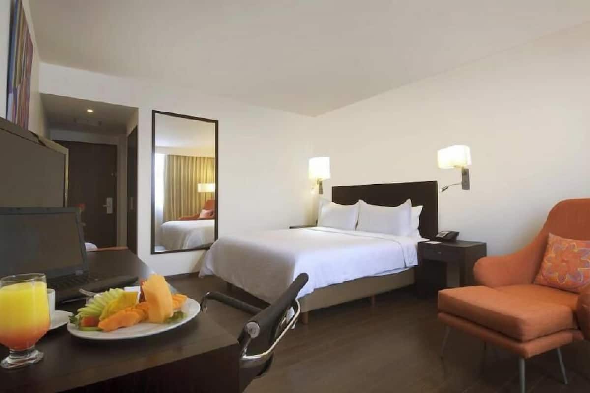 A hotel room with a bed, bedside table, chair, and a desk with a laptop and fruit.