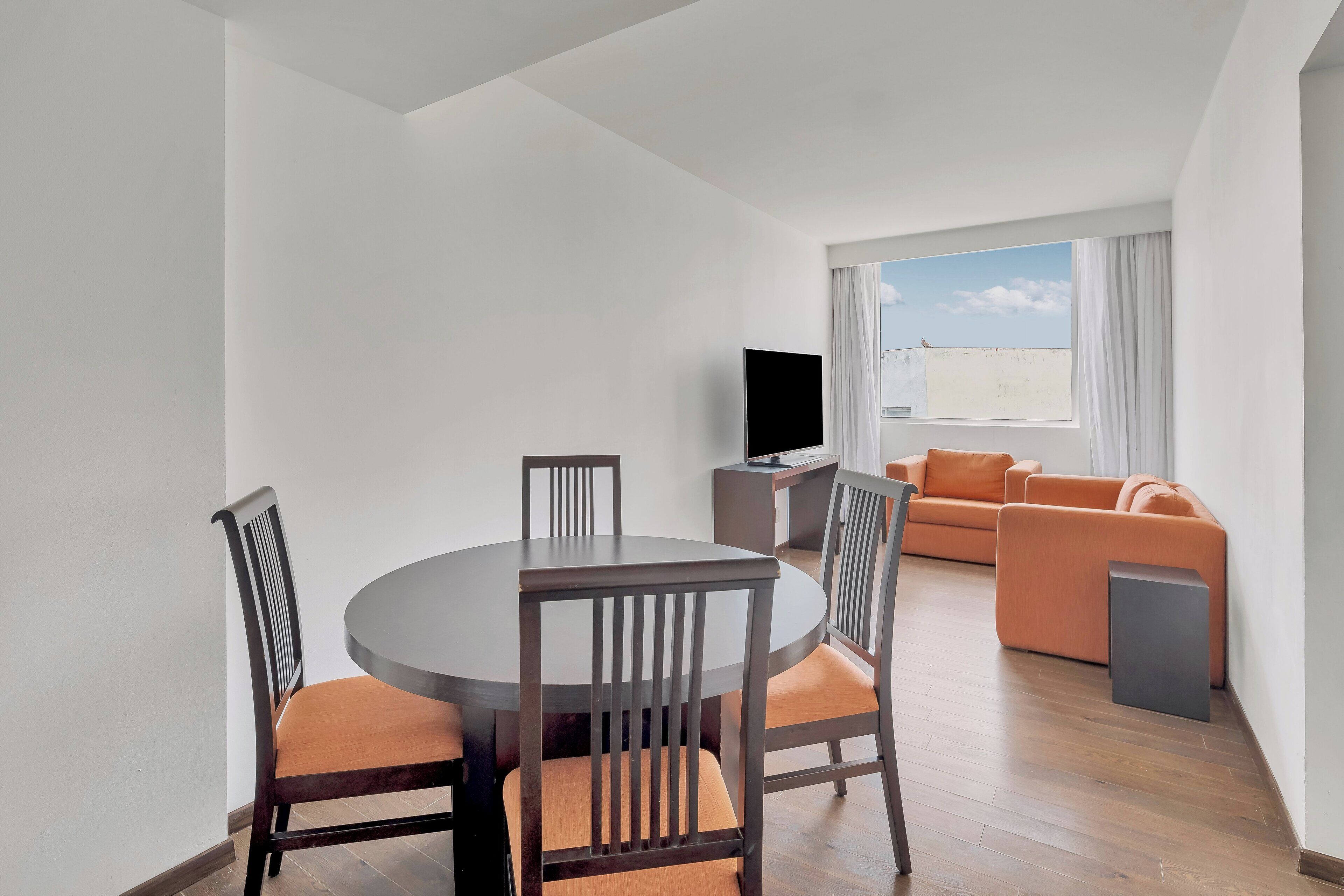A modern living room with a round dining table, wooden chairs, a sofa, and a flat-screen TV.