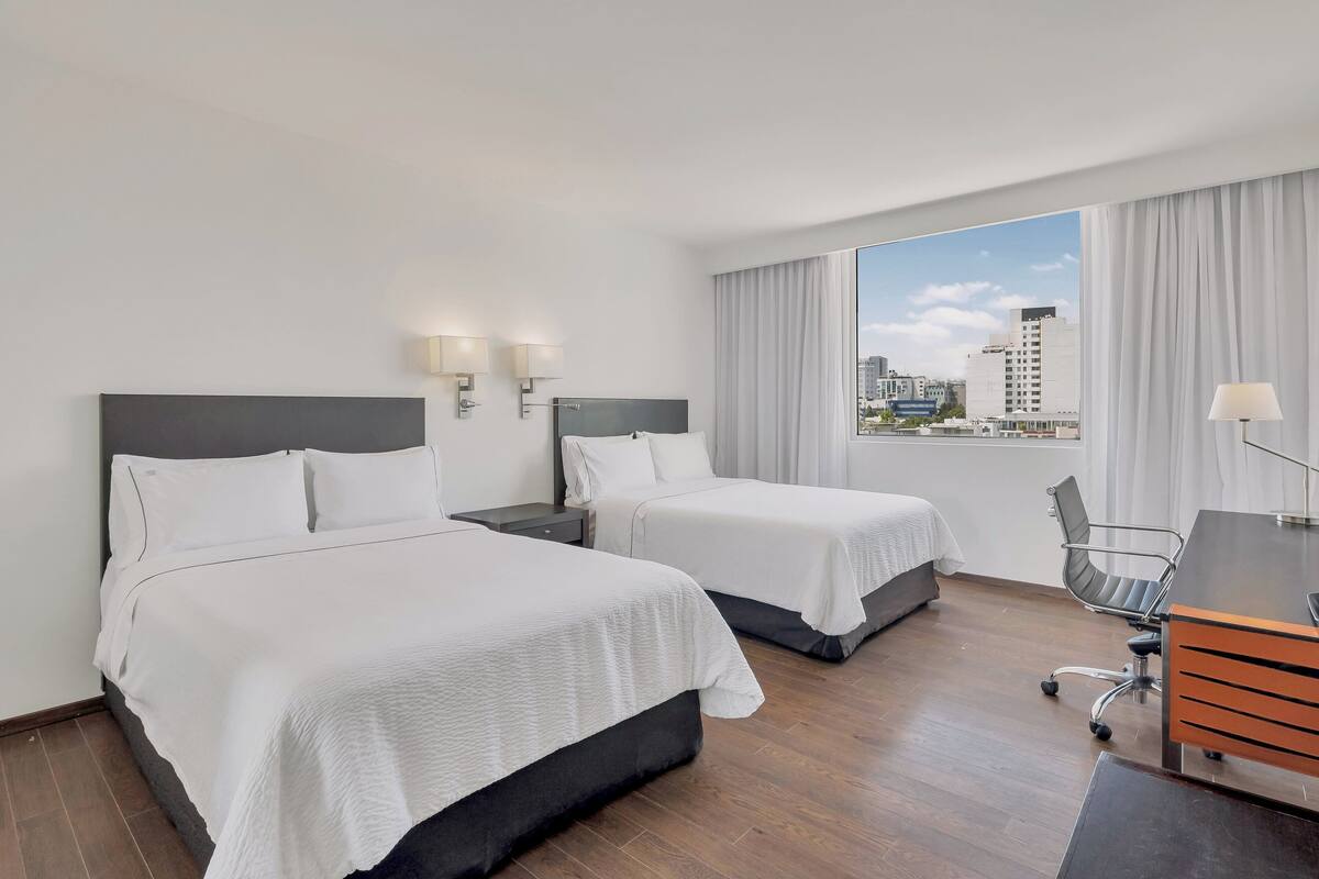 A hotel room with two beds, a desk, and a view of the city.
