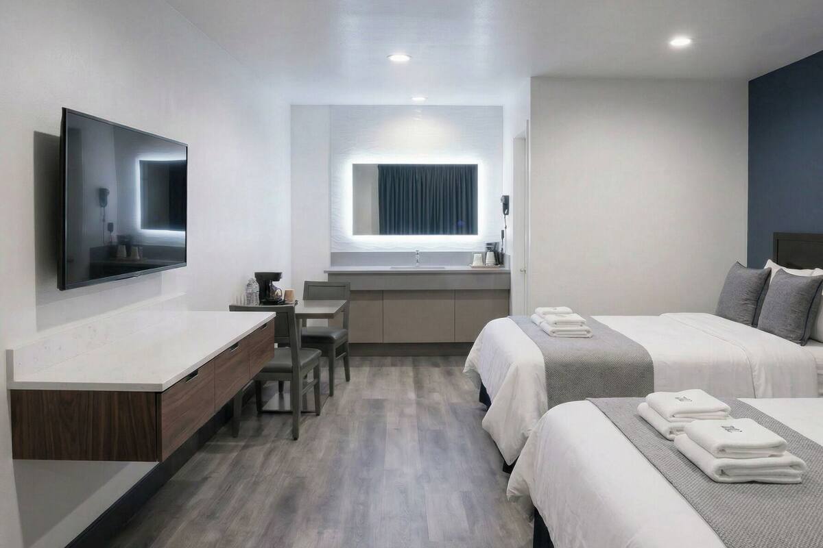 A modern hotel room with a flat-screen TV, a desk with a chair, a large window, two beds with towels, and a wooden floor.