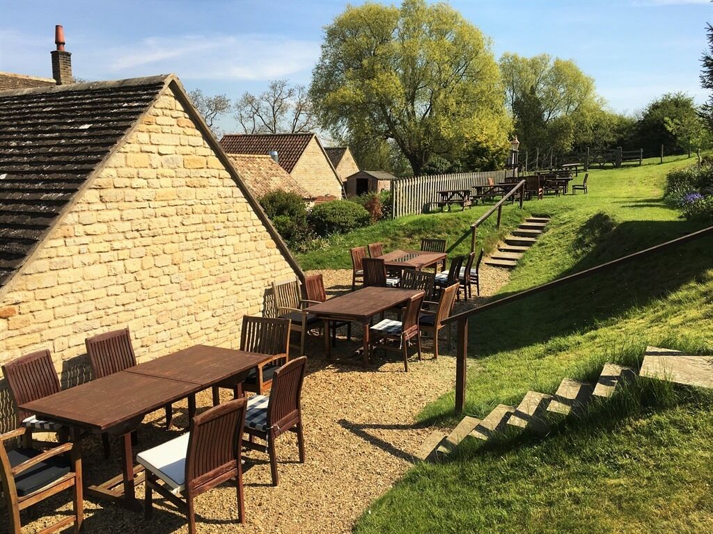 Un espace extérieur aménagé dans un style rustique, avec des tables et des chaises en bois, entouré d’un bâtiment en pierre et d’une pente herbeuse.