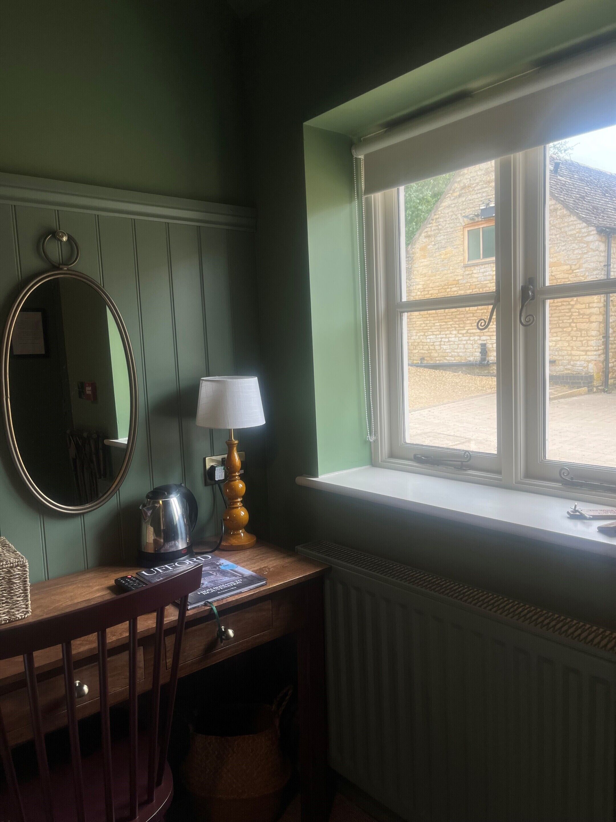 Una habitación con un escritorio de madera, un espejo, una lámpara, un radiador y una ventana que ofrece vistas a un edificio de piedra.