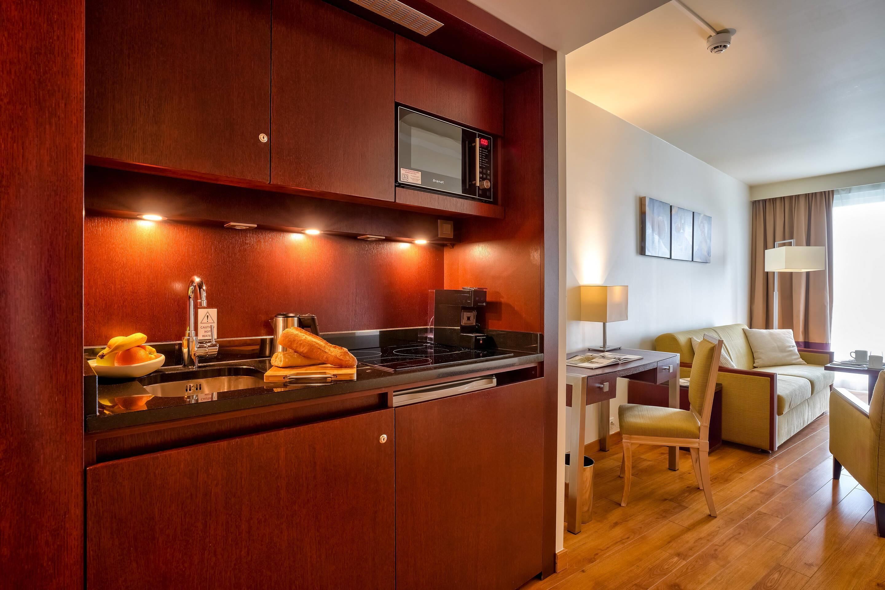 A compact hotel room with a kitchenette, a bed, a desk, and a chair.