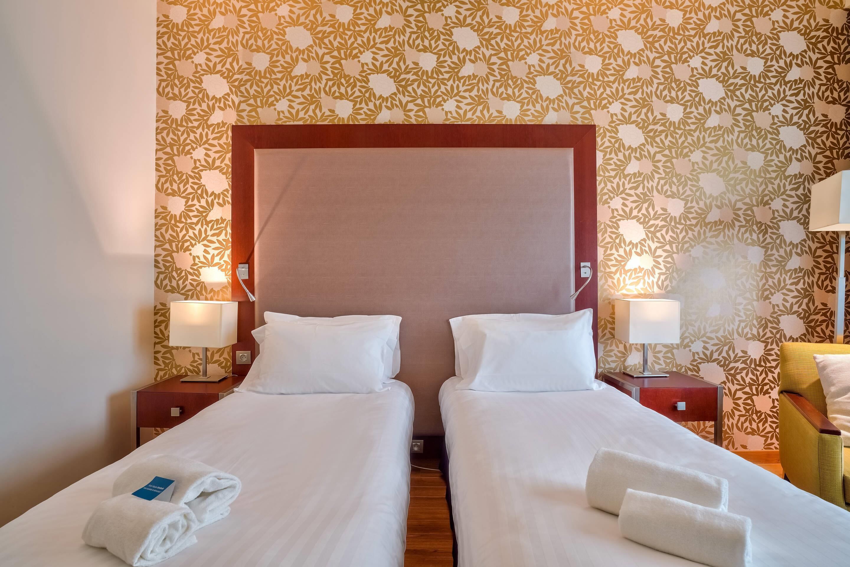 Une chambre d’hôtel avec deux lits simples, une tête de lit en bois, un papier peint à motifs floraux, des lampes de chevet et un fauteuil jaune.