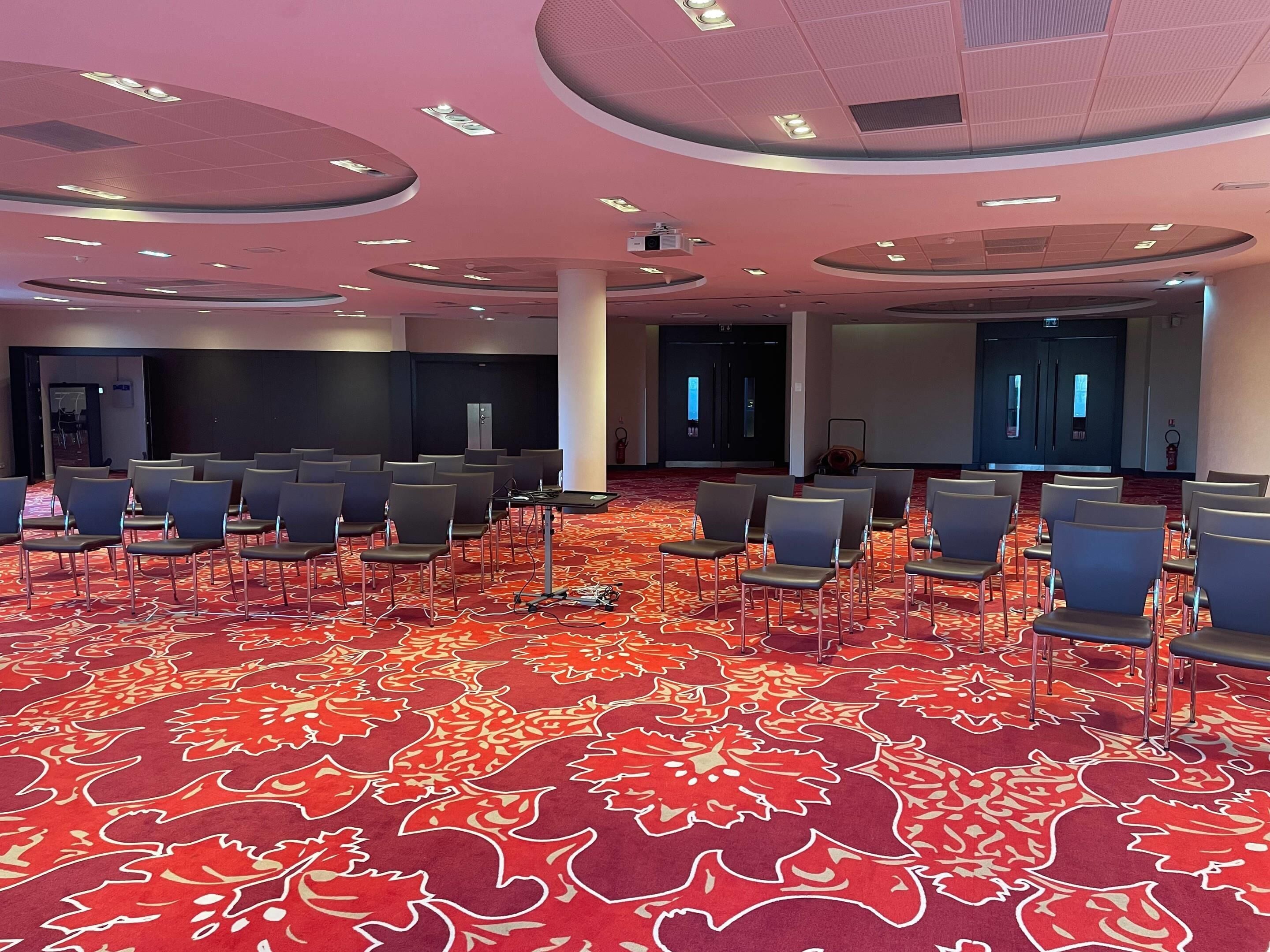 Ein Konferenzraum mit Reihen von Stühlen, einem roten Teppich mit Muster und einem modernen Deckenstil.