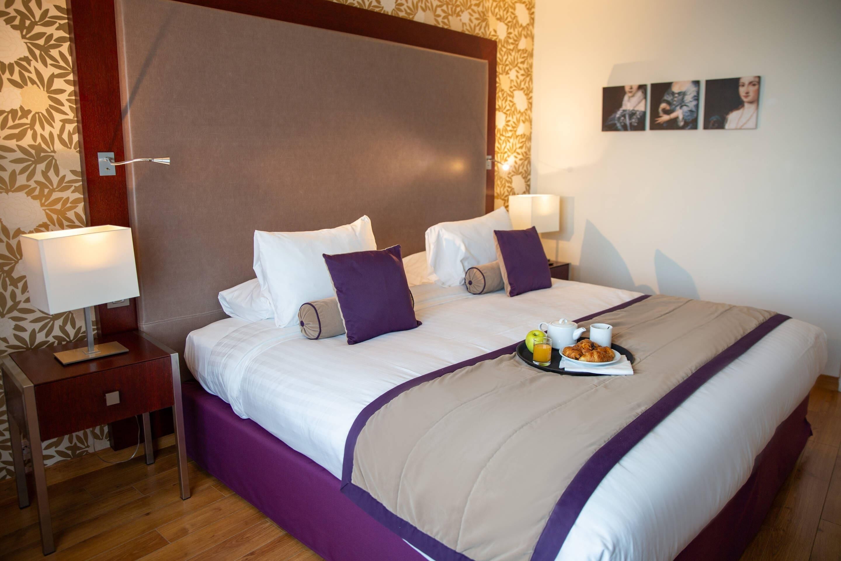 Une chambre d’hôtel avec un grand lit, deux tables de chevet, une petite table et un plateau contenant des articles pour le petit-déjeuner.