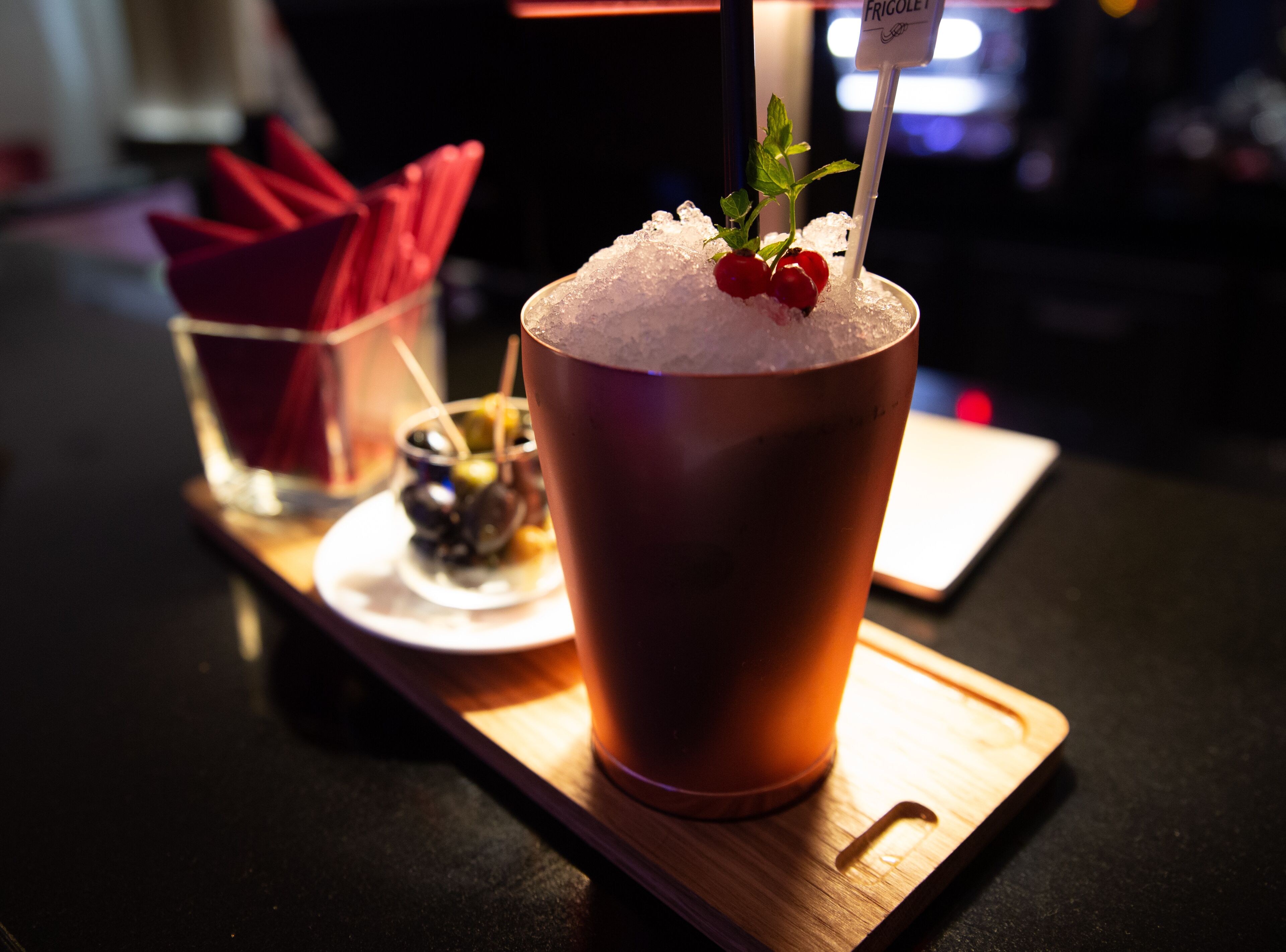 Ein Cocktail mit Minze als Garnitur und einem Strohhalm, serviert auf einem Holztablett mit einer Serviette und einer kleinen Oliven-Schale.