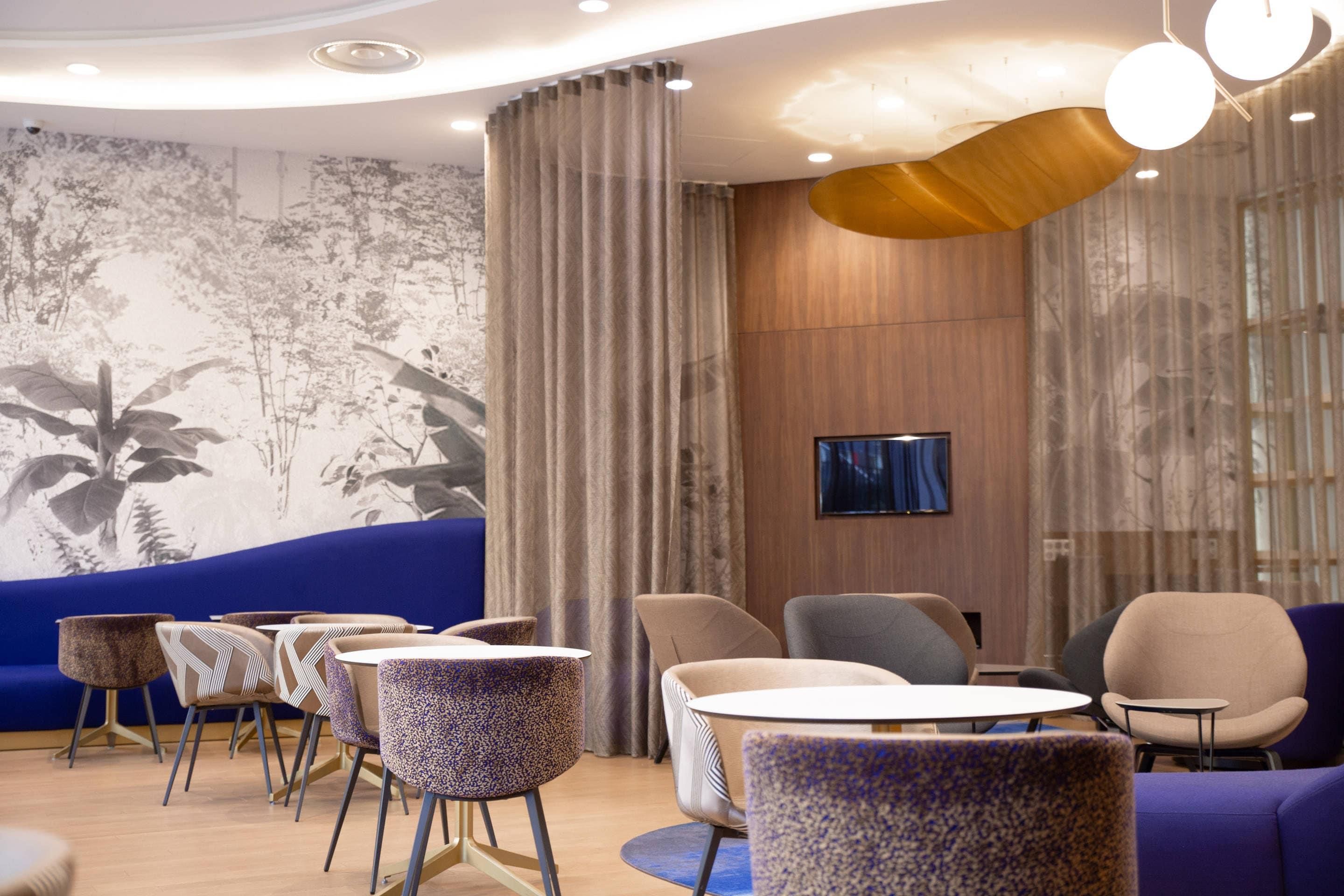 Ein modernes Interieur mit einem blauen Sofa, runden Tischen, Stühlen, einem an der Wand montierten Fernseher und einem Wandgemälde mit Muster.