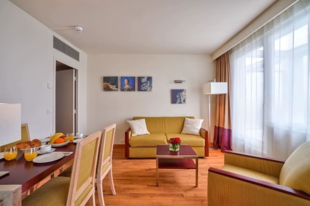 A hotel room with a dining area, a sofa, a coffee table, and artwork on the walls.