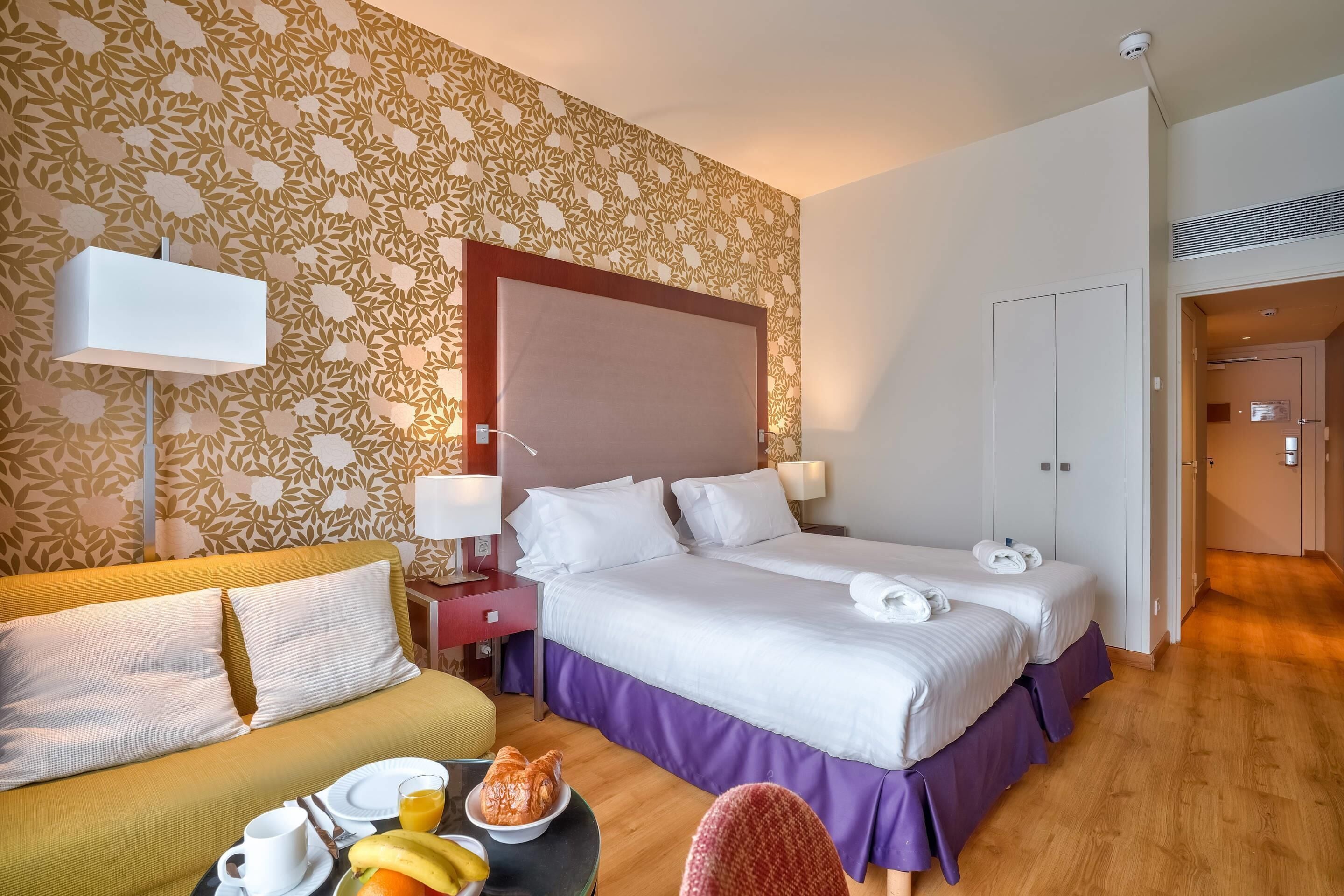 A hotel room with a bed, a sofa, a dining table with breakfast, and a patterned wallpaper.