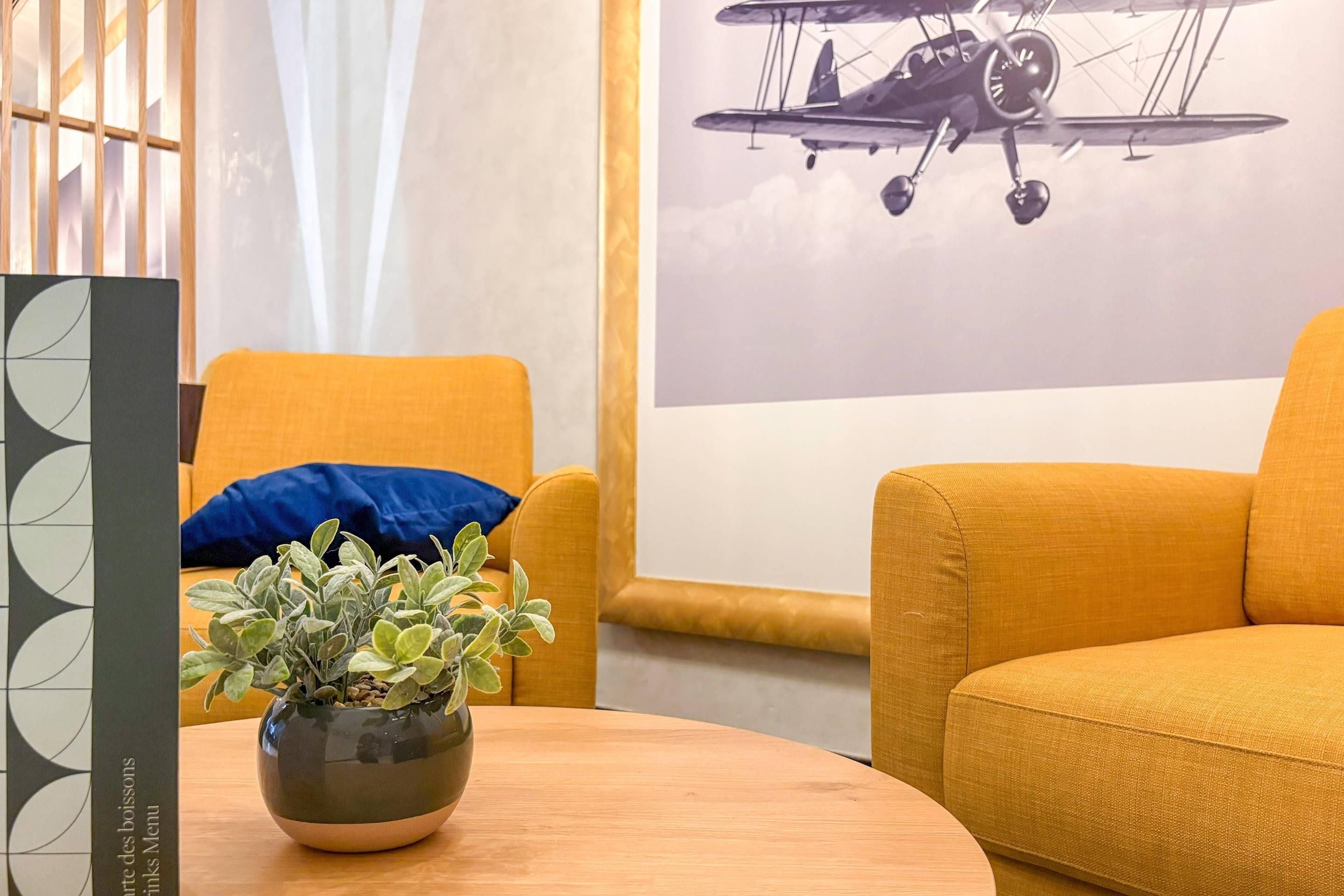 Een kamer met een ronde houten tafel, een plant in pot, twee gele fauteuils, een blauw kussen en een foto van een vliegtuig aan de muur.