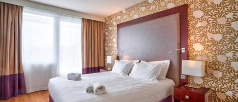 Quarto de hotel com uma cama grande, mesa de cabeceira, luminária e janela com cortinas.
