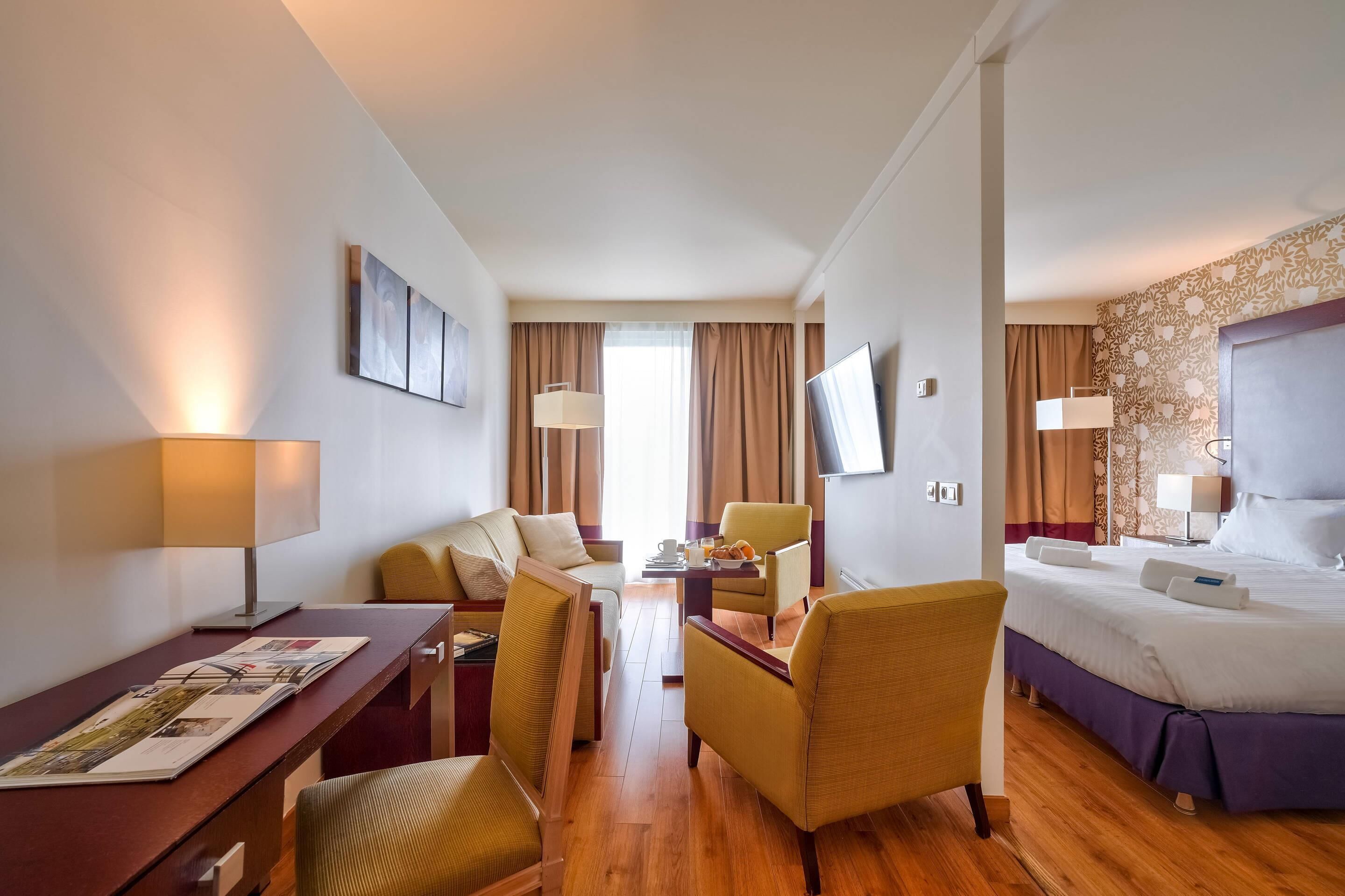 Una habitación de hotel con cama, escritorio con lámpara, dos sillas, televisor y ventana con cortinas.