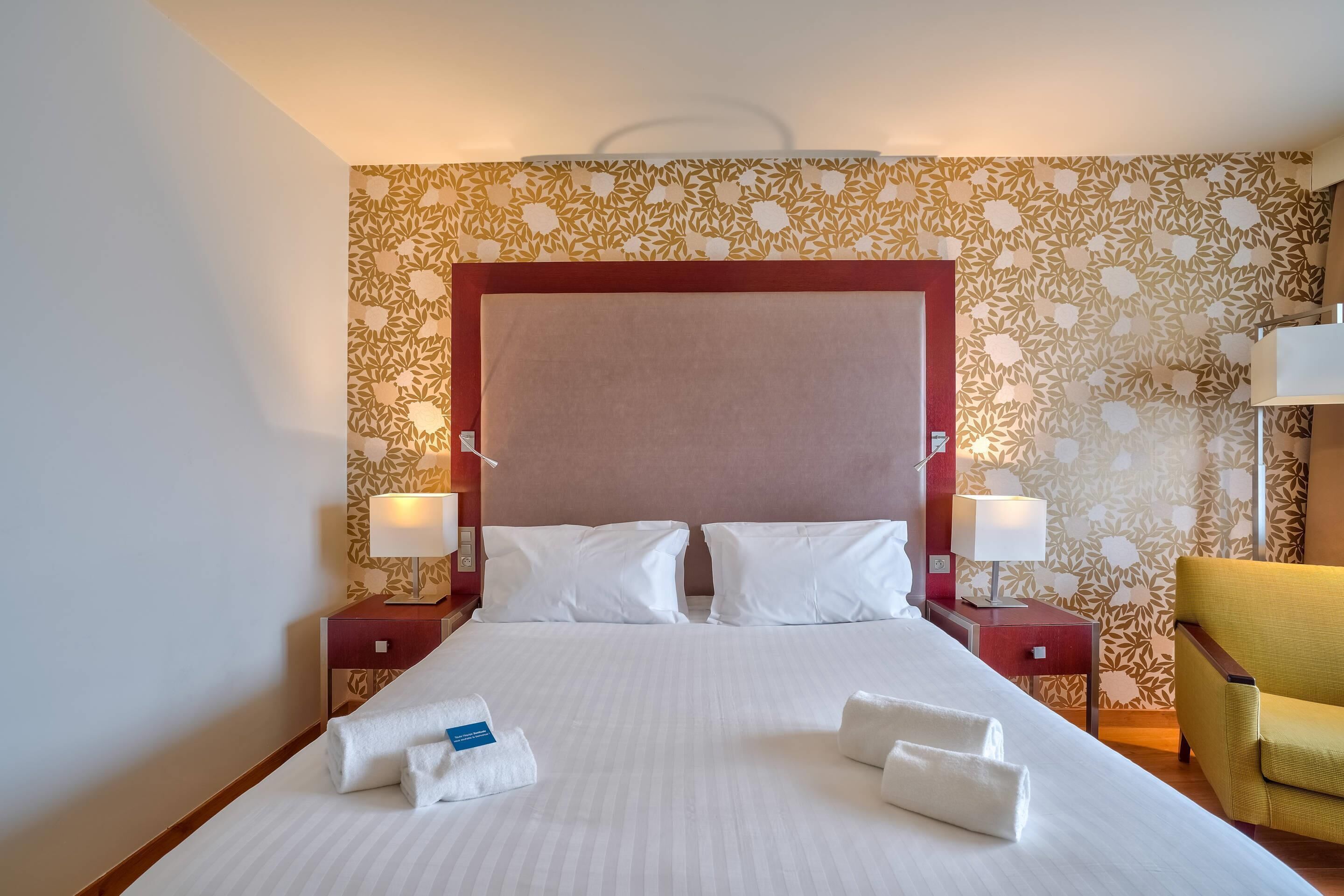 Una camera d'albergo con un letto grande, due comodini con lampade, una poltrona gialla e carta da parati con motivi floreali.
