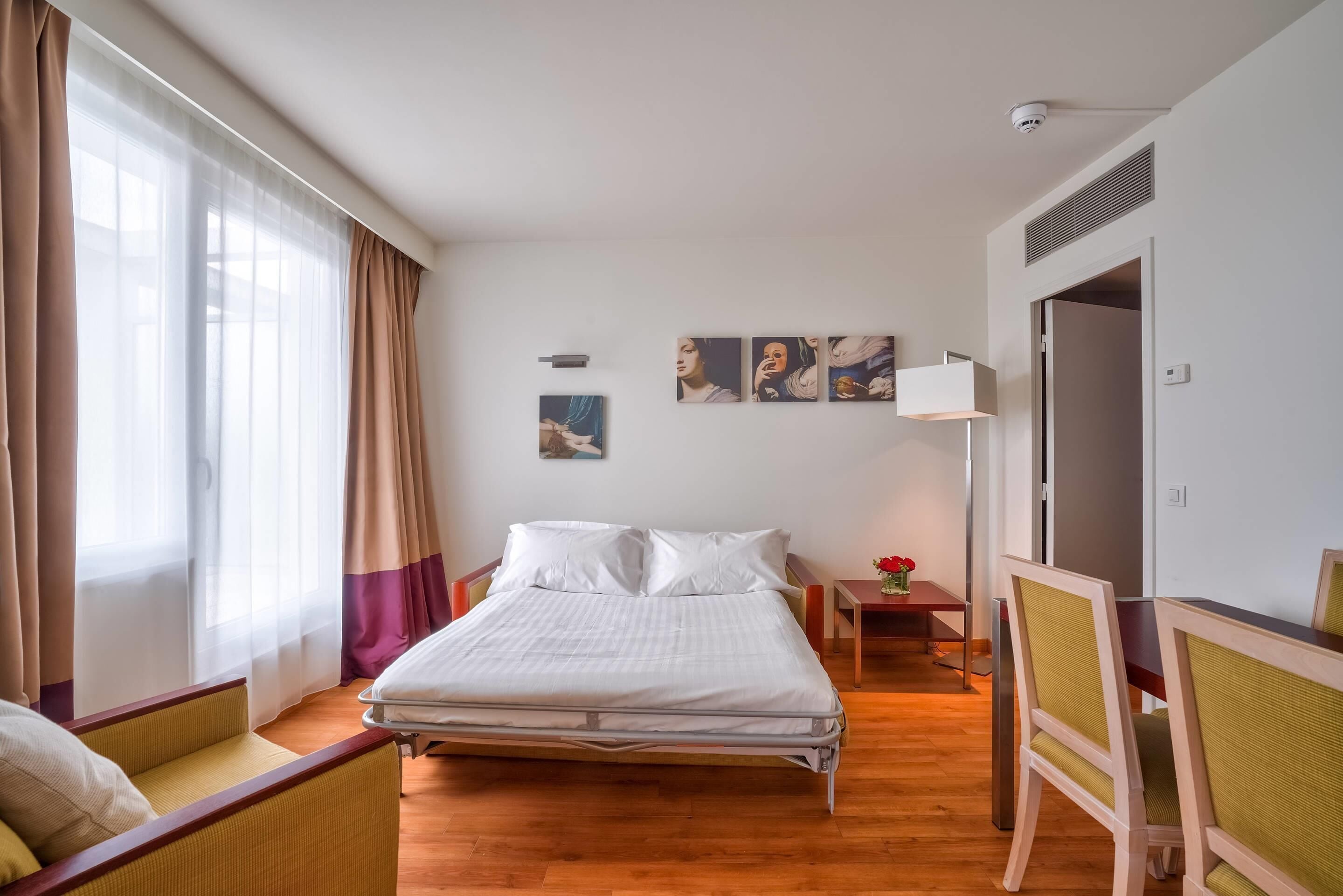 Ein Hotelzimmer mit Bett, Sessel, kleinem Tisch mit Blumenvase und einem Gemälde an der Wand.