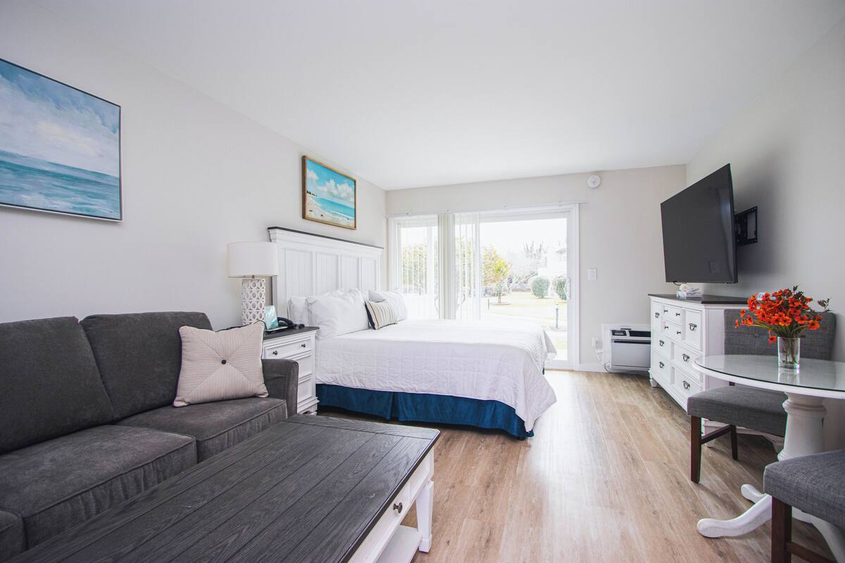 A bedroom with a bed, a grey sofa, a bedside table, a TV, and a window.