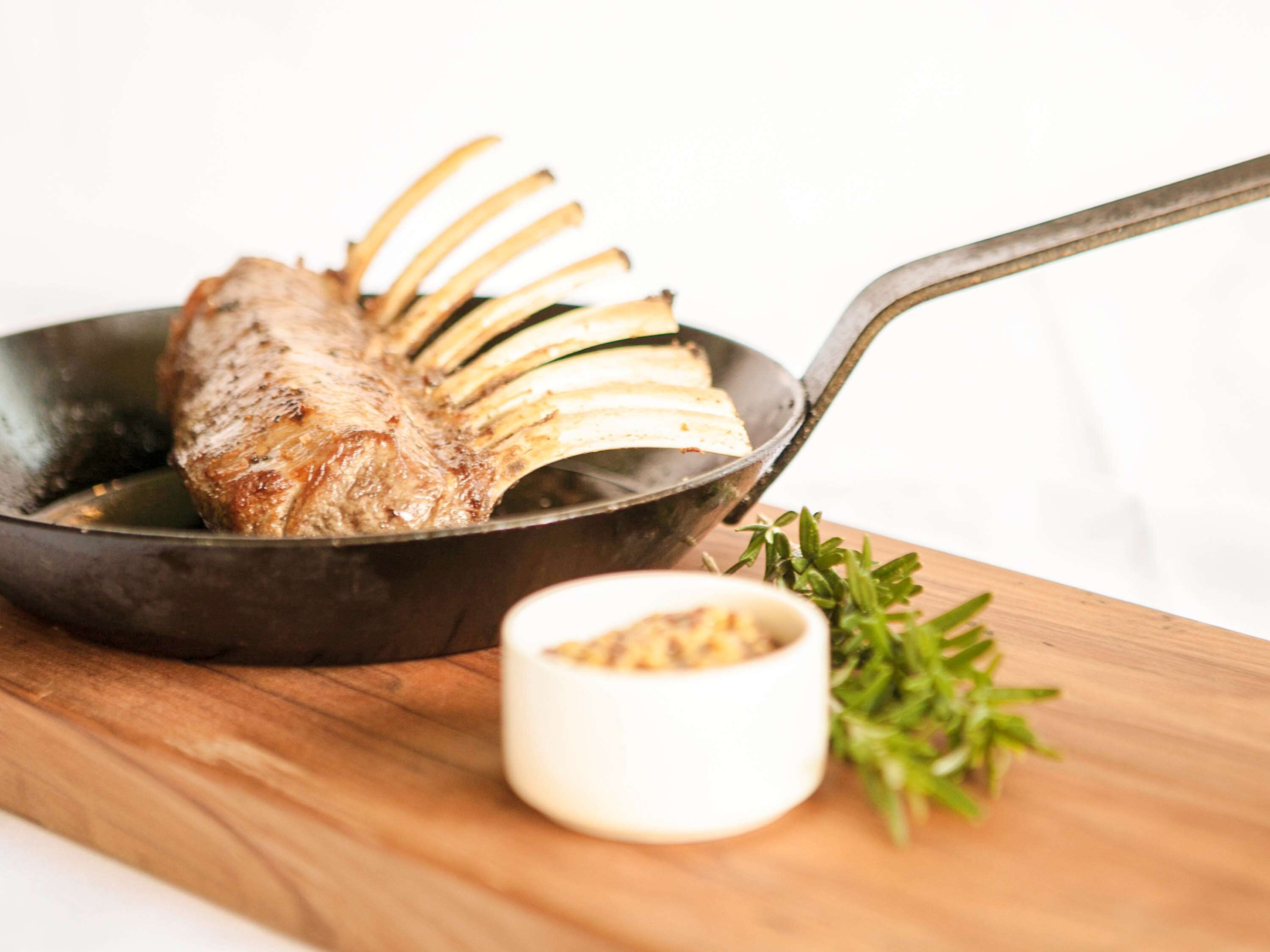 A cooked rack of lamb in a pan with a handle, accompanied by a small bowl of seasoning and a sprig of rosemary on a wooden board.