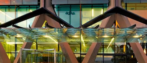 A modern hotel entrance with a glass facade, geometric architectural design, and a visible reception area.