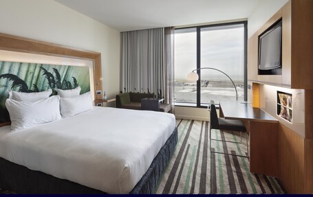 A hotel room with a large bed, a desk, a chair, a TV, and a window with a city view.