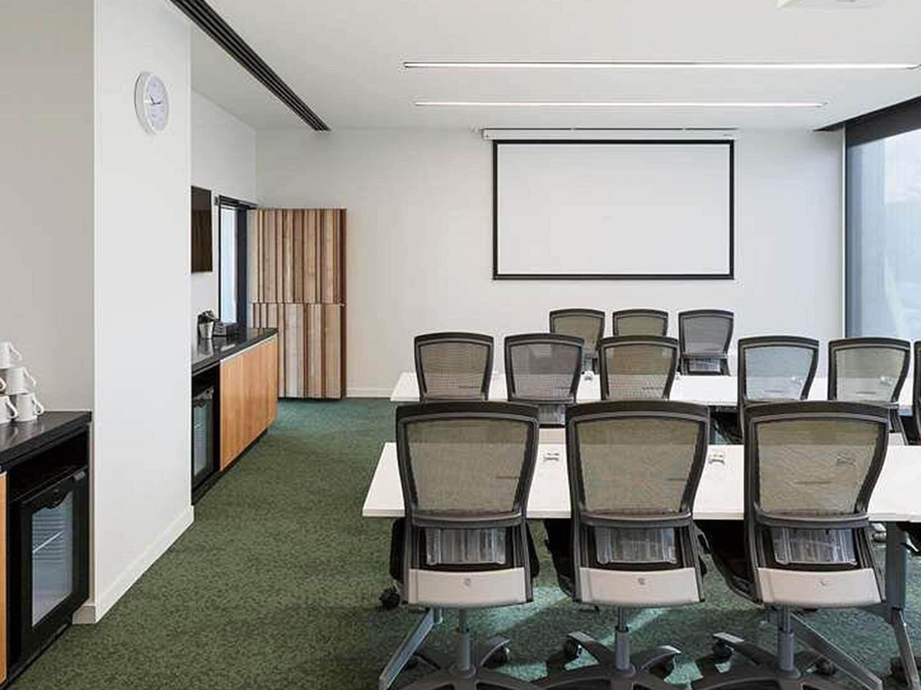 A conference room with a large table, chairs, a projector screen, a clock, and a window.