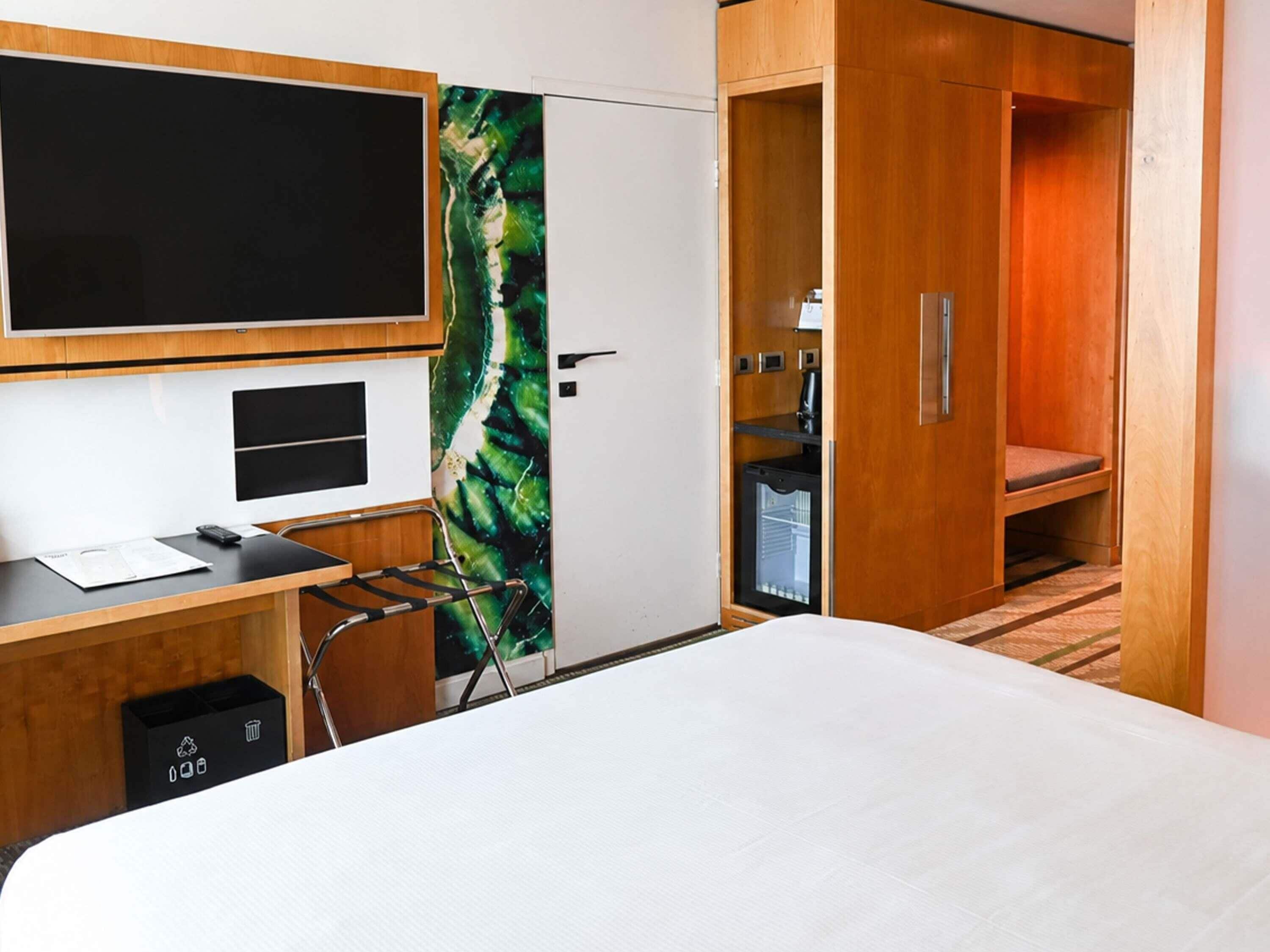 A hotel room with a bed, a desk with a computer, a TV, a wardrobe, and a small kitchenette.