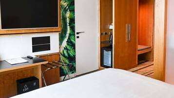 A hotel room with a bed, a desk with a computer, a TV, a wardrobe, and a small kitchenette.