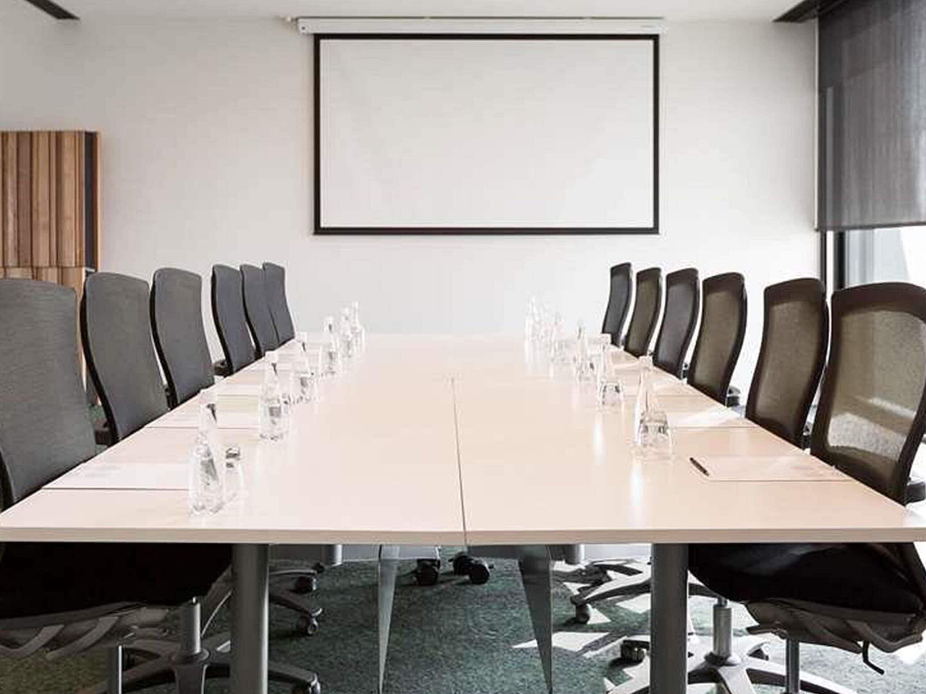 A conference room with a long table, chairs, a projector screen, and a carpeted floor.