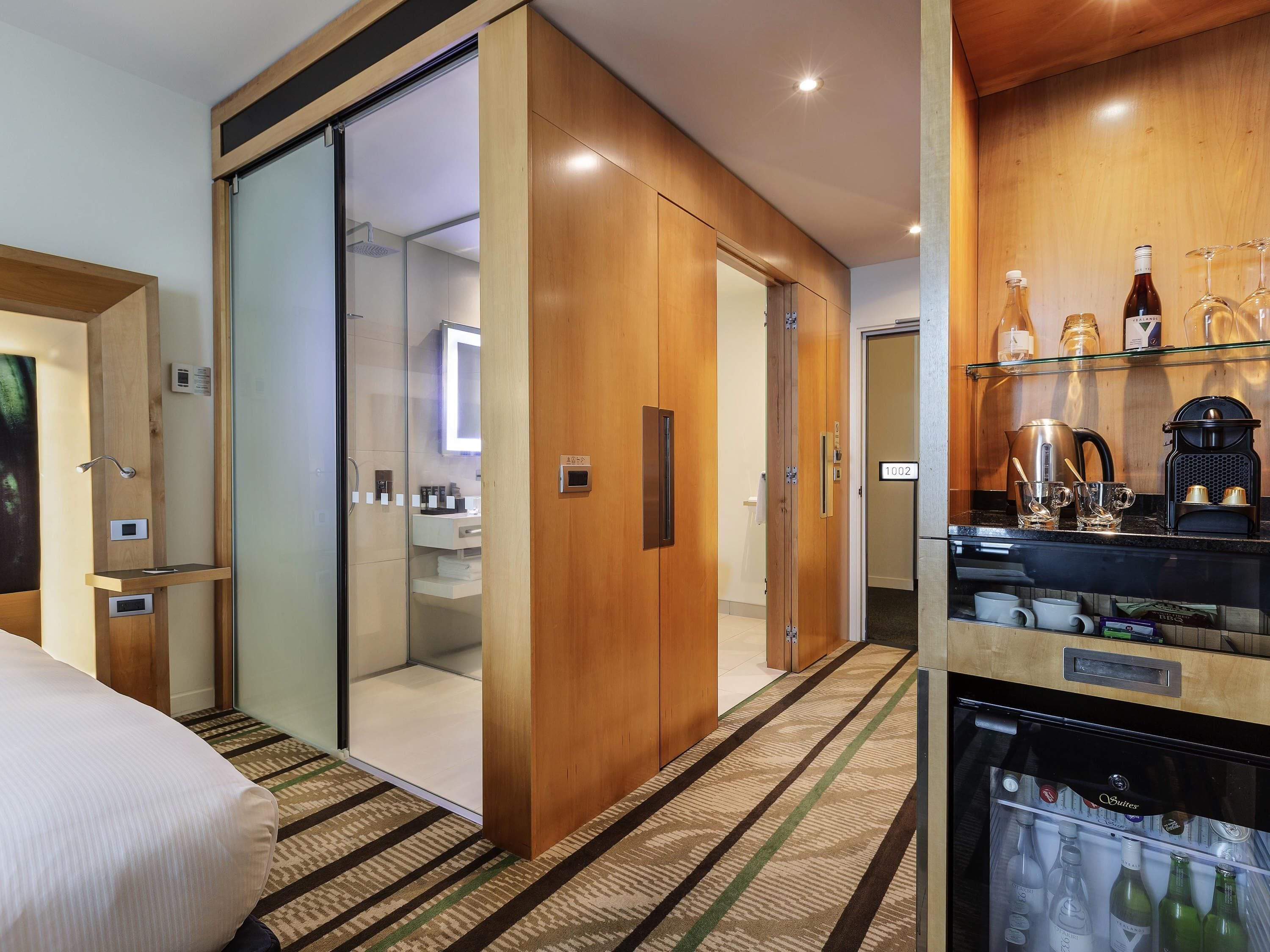 Habitación de hotel con puerta corredera que da a un baño, un minibár con bebidas y una cafetera.