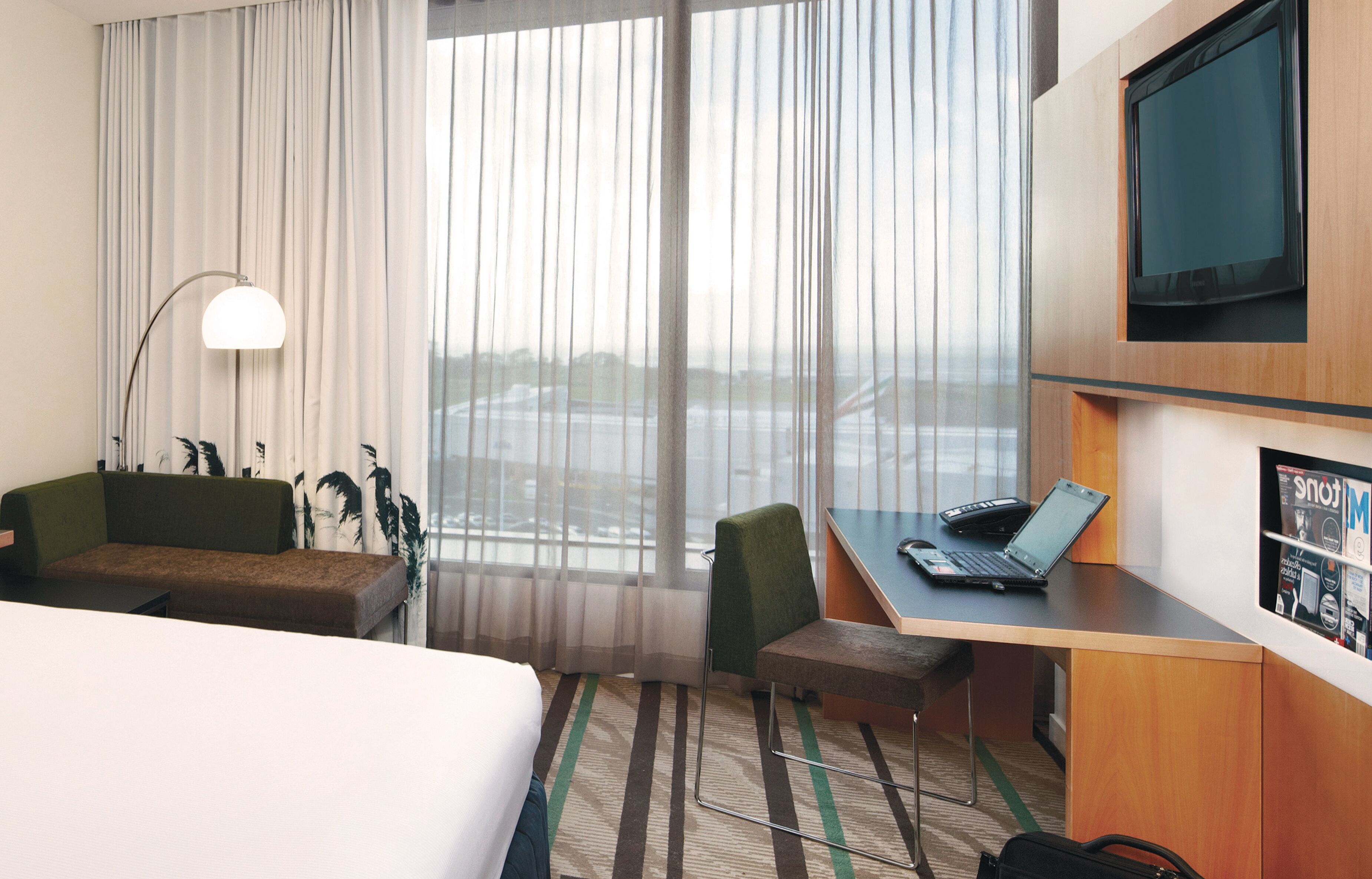 A hotel room with a bed, a desk, a chair, a lamp, a TV, and a view of the city through large windows.