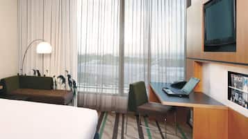 A hotel room with a bed, a desk, a chair, a lamp, a TV, and a view of the city through large windows.