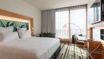A hotel room with a large bed, a desk, a chair, a TV, and a view of the city through the window.