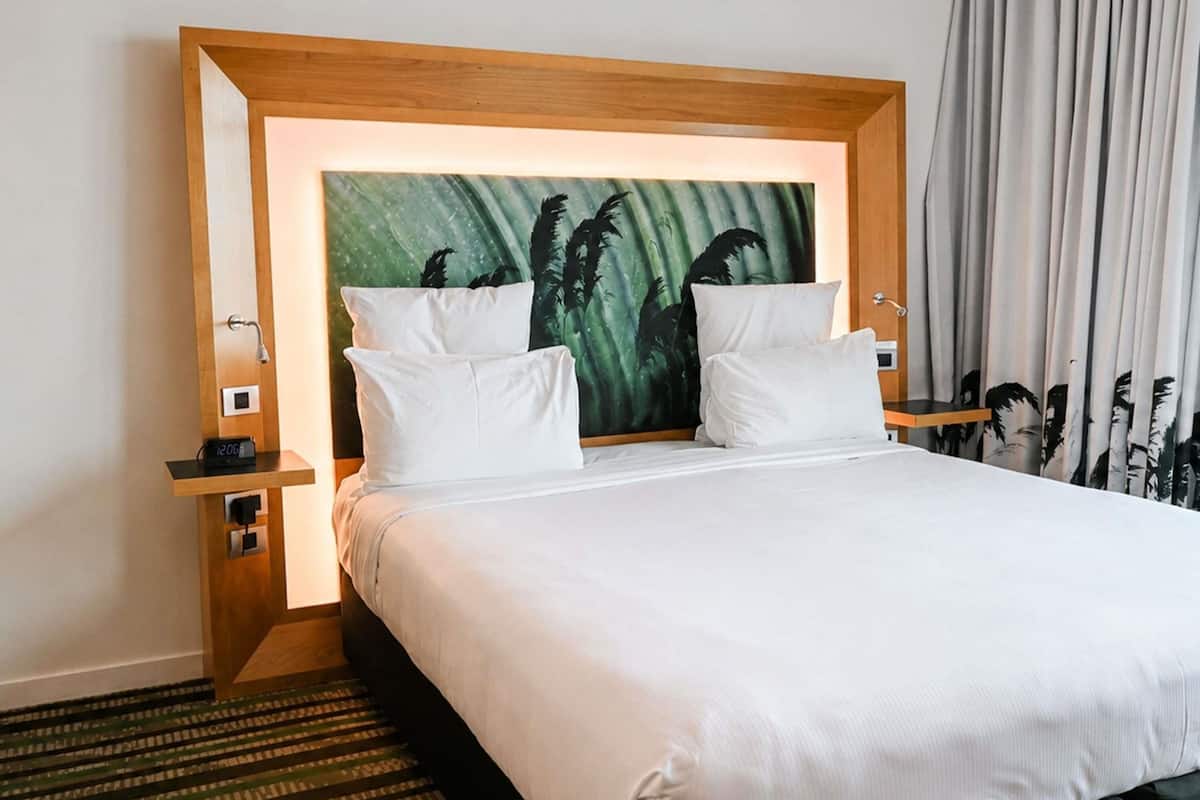 A hotel room with a large bed, two white pillows, a headboard with a tropical painting, a nightstand with a clock, and a window with curtains.
