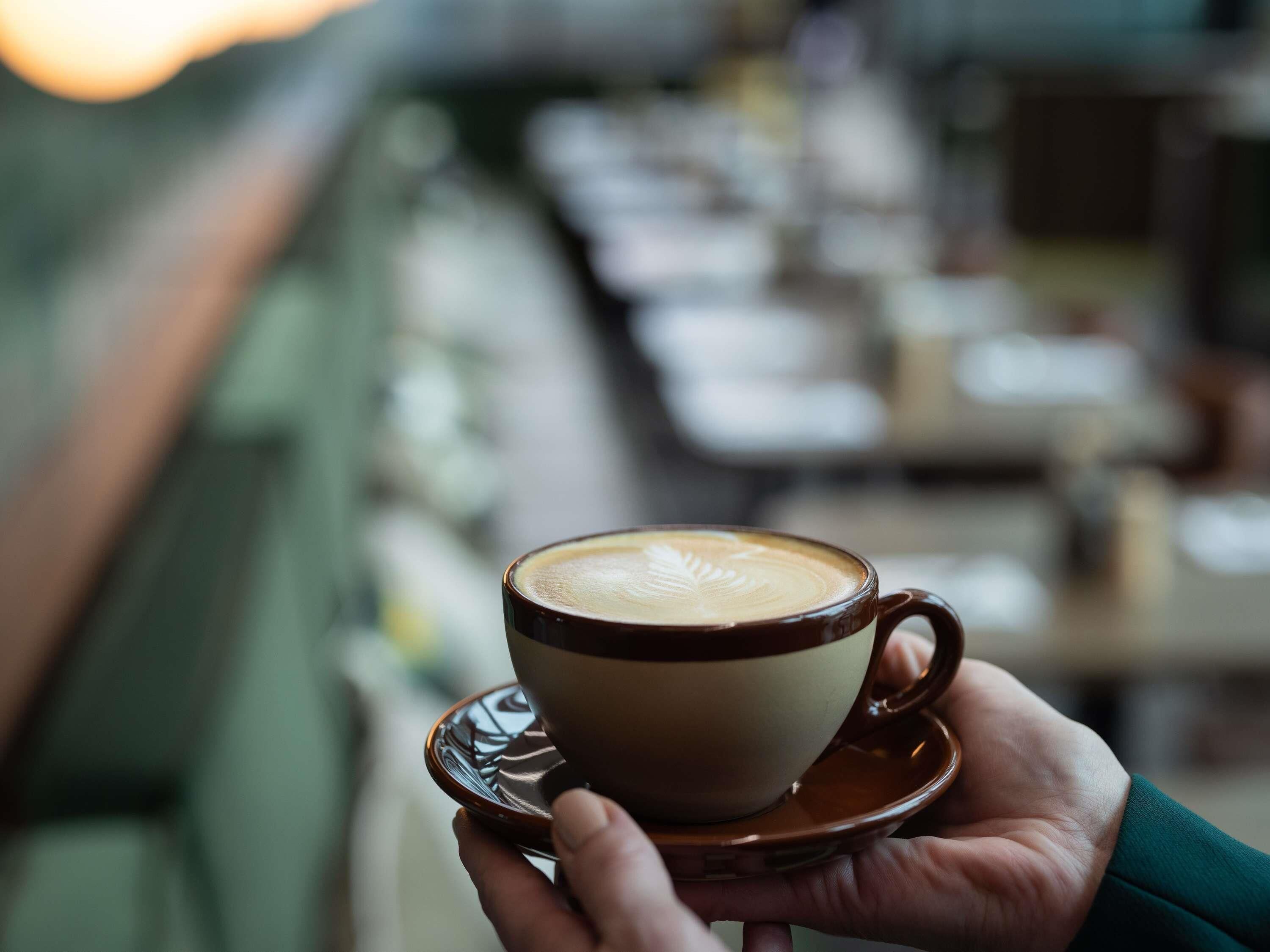 A hand holding a cup of coffee with a saucer, in a blurred indoor setting.