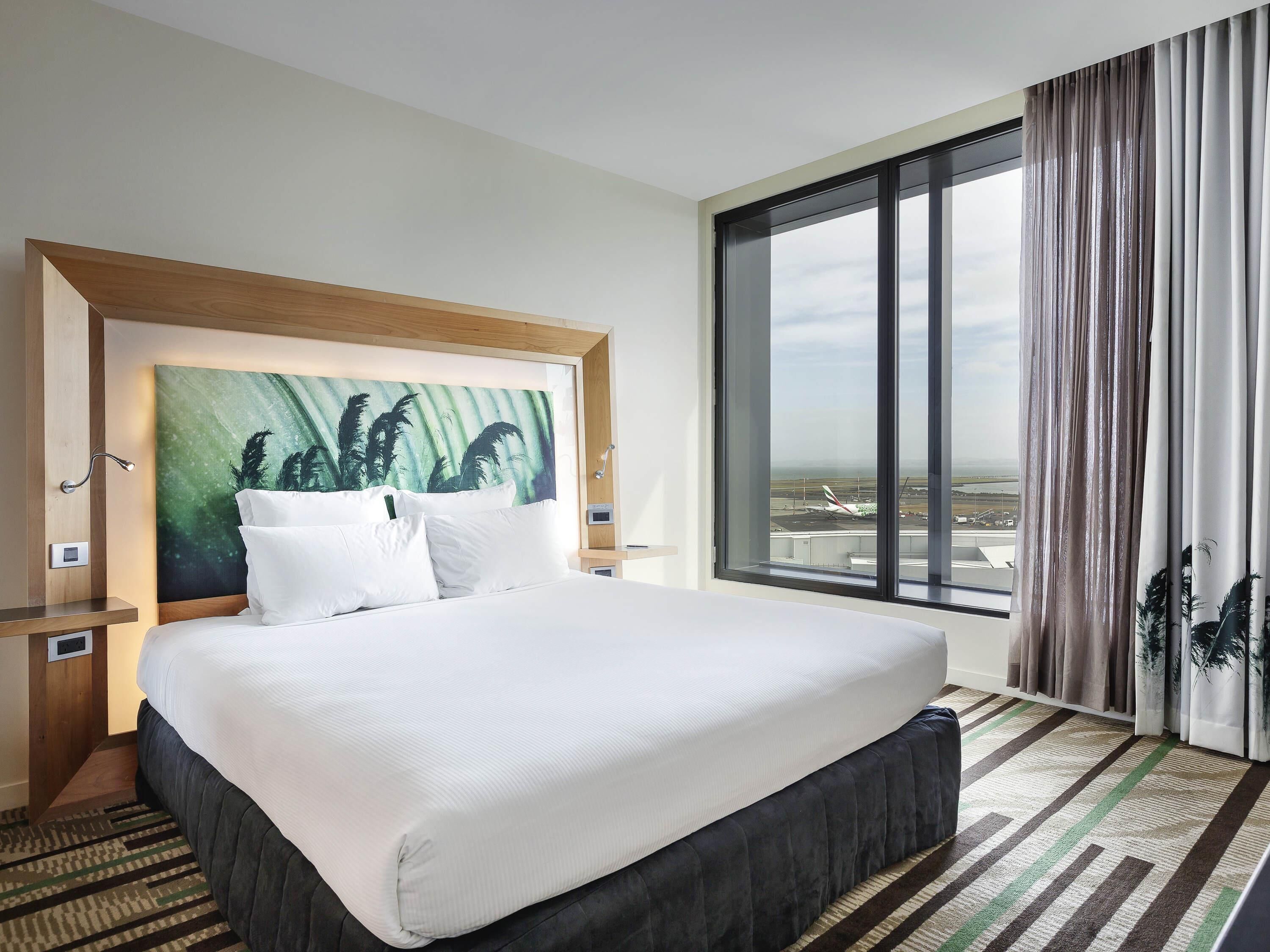 A hotel room with a large bed, a headboard with a green abstract painting, a view of an airport, and a striped carpet.