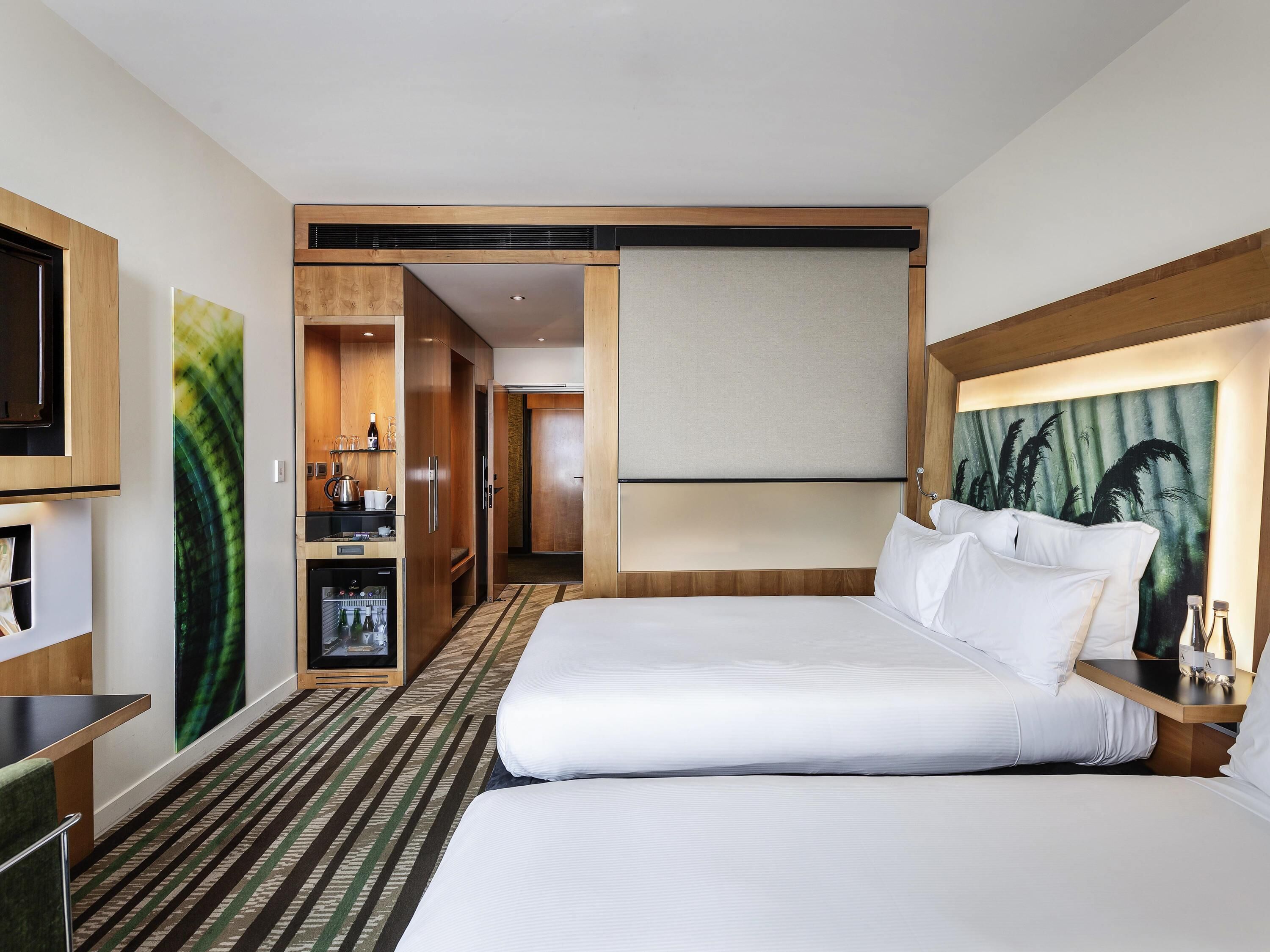 A hotel room with a bed, a desk, a minibar, a television, and a painting on the wall.