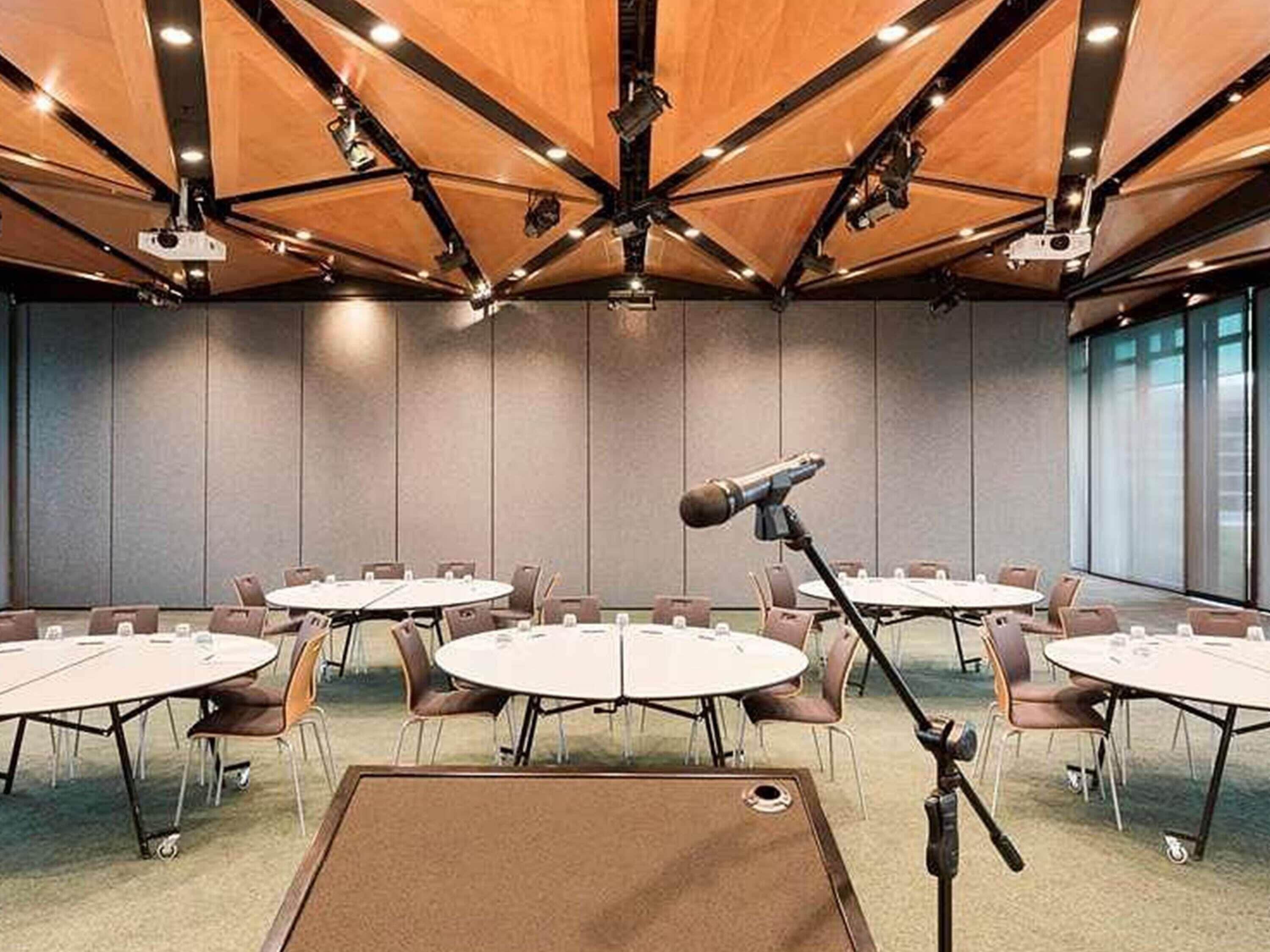 Una sala conferenze con più tavoli, sedie, un leggio e un soffitto con travi e illuminazione a vista.