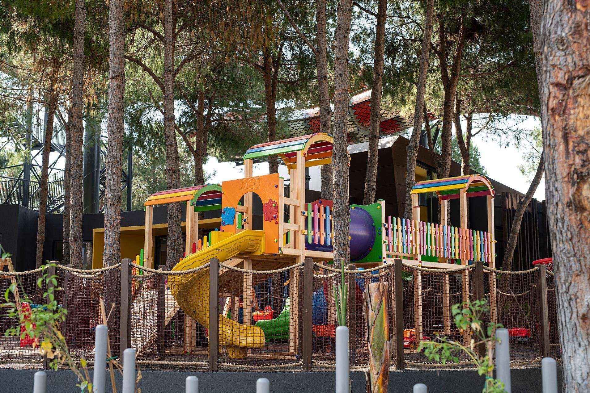 Un parque infantil con estructura de madera que incluye toboganes y zonas de escalada, rodeado de árboles y una valla.