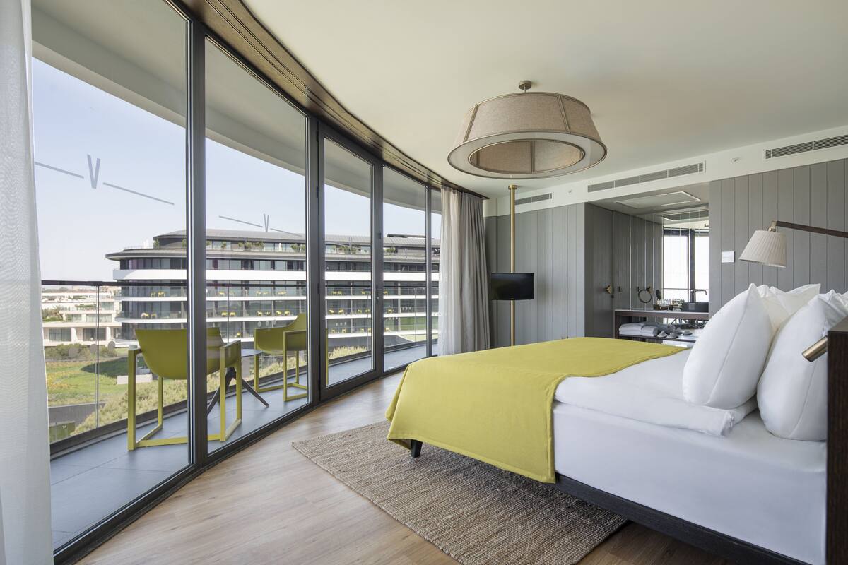 A modern hotel room with a large bed, a view of a cityscape, and a balcony with chairs.