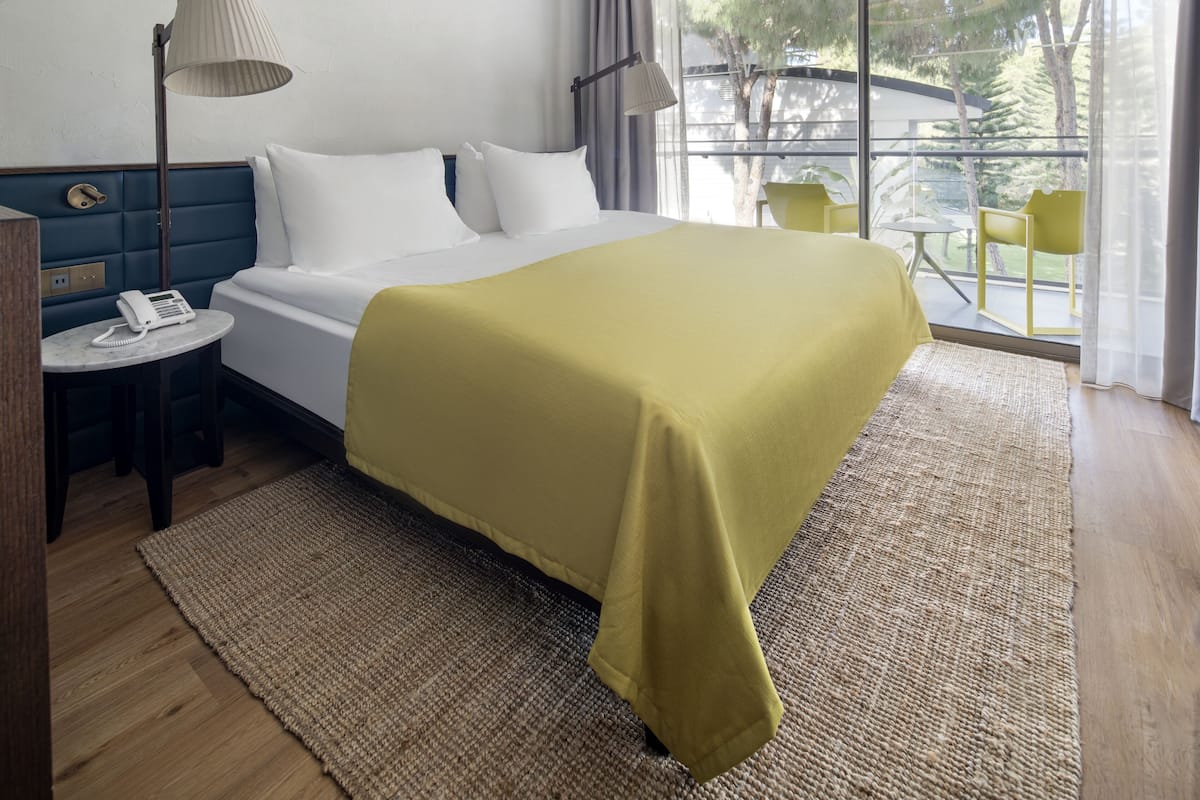 A hotel room with a bed, bedside lamps, a telephone, a small table, and a view of the outdoors through large windows.