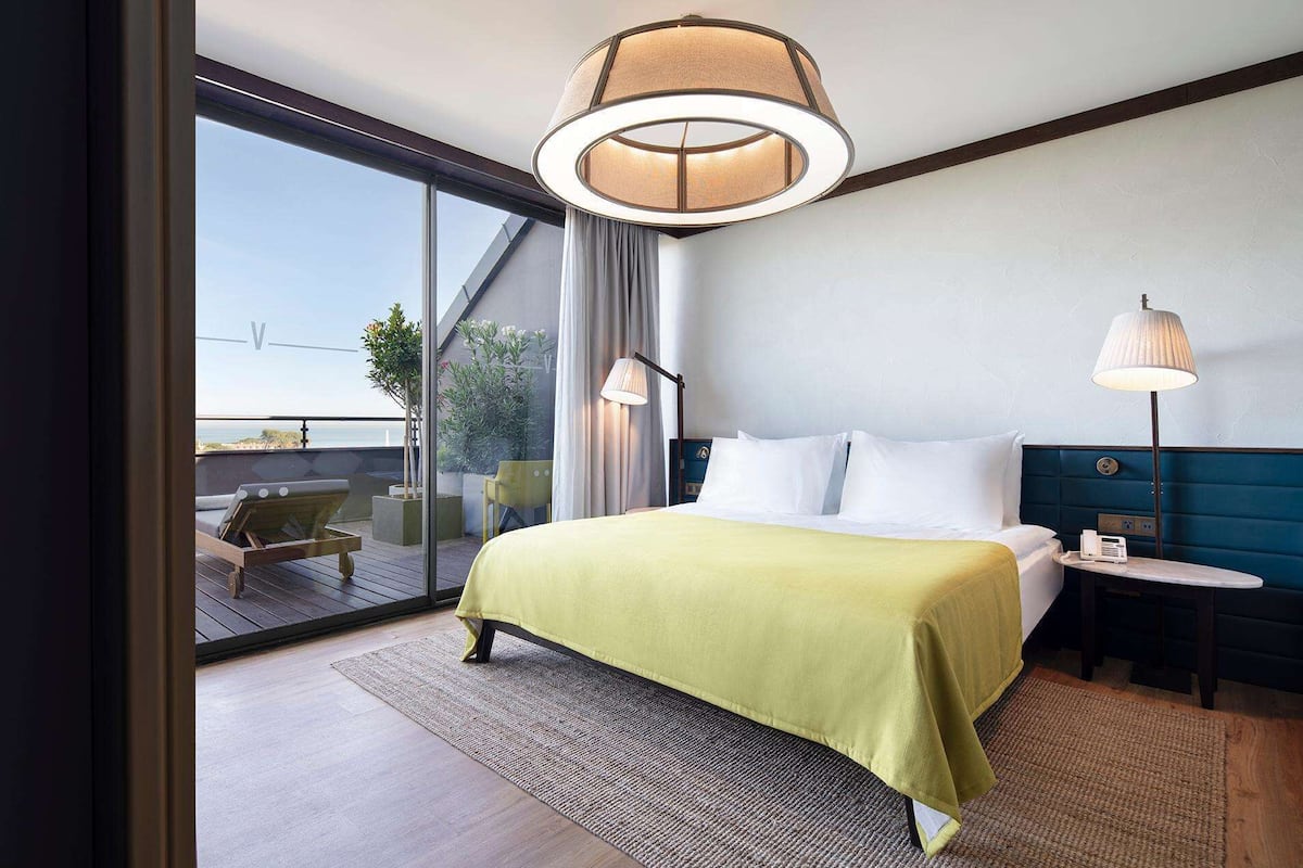 A hotel room with a bed, bedside table, lamp, and a view of the outdoors.