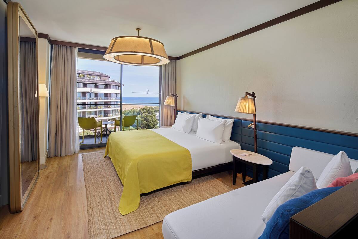 A hotel room with a large bed, a smaller bed, a desk, and a view of a building and greenery outside.