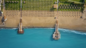 An aerial view of a beach resort with two wooden piers extending into the sea, each with rows of round tables and chairs. - Voyage Belek Golf & Spa (Serik)