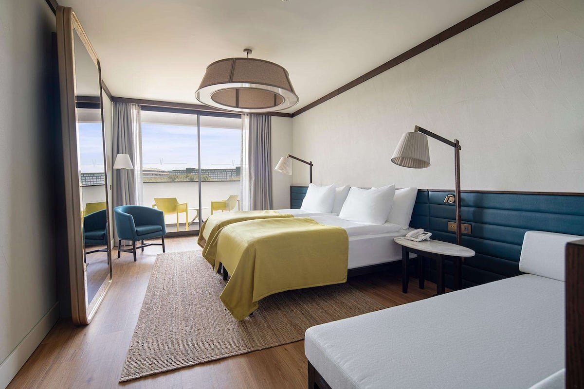 A hotel room with a bed, bedside table, lamp, and a view of the outdoors.