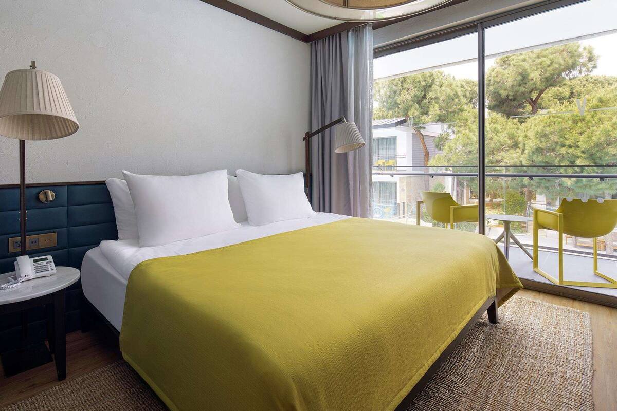 A hotel room with a bed, a bedside table with a radio, a large window with a view of trees, and a balcony with yellow chairs.