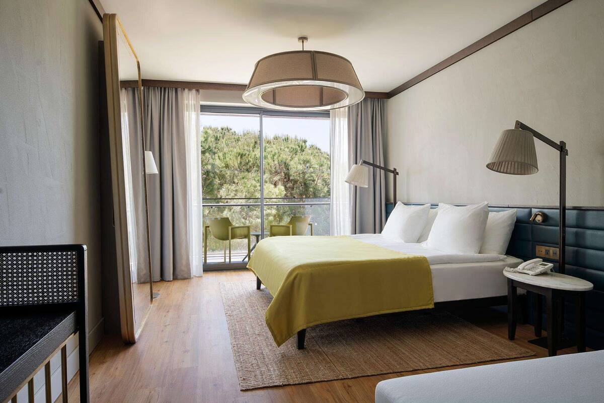 A hotel room with a bed, bedside table, lamp, and a view of greenery through a large window.
