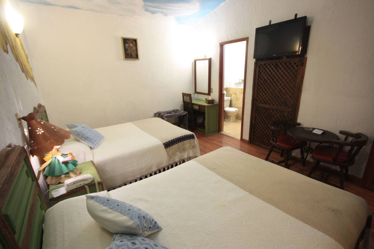 Quarto de hotel com duas camas, uma escrivaninha, uma cadeira, uma televisão e um banheiro visível através de uma porta aberta.