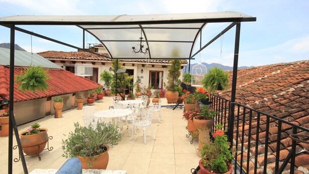 A covered patio area with a table and chairs, surrounded by potted plants and a view of a building with a tiled roof.